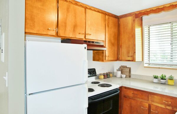 Rendering of a kitchen with wood cabinets, gray countertops, a white fridge, and a window.