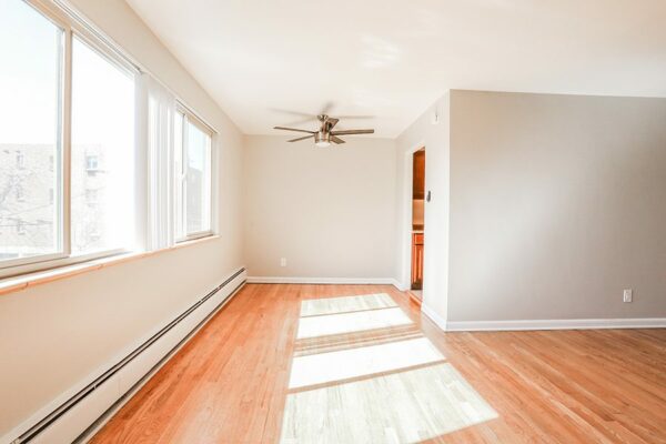 Living room with wood floor, warm grey walls, baseboard heaters, ceiling fan, and large windows.