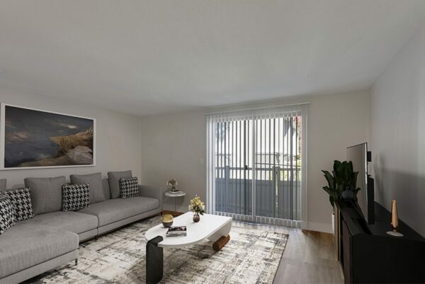 Living room with wood floors covered with a large carpet, a gray plush couch, and white walls with a door leading to a patio.